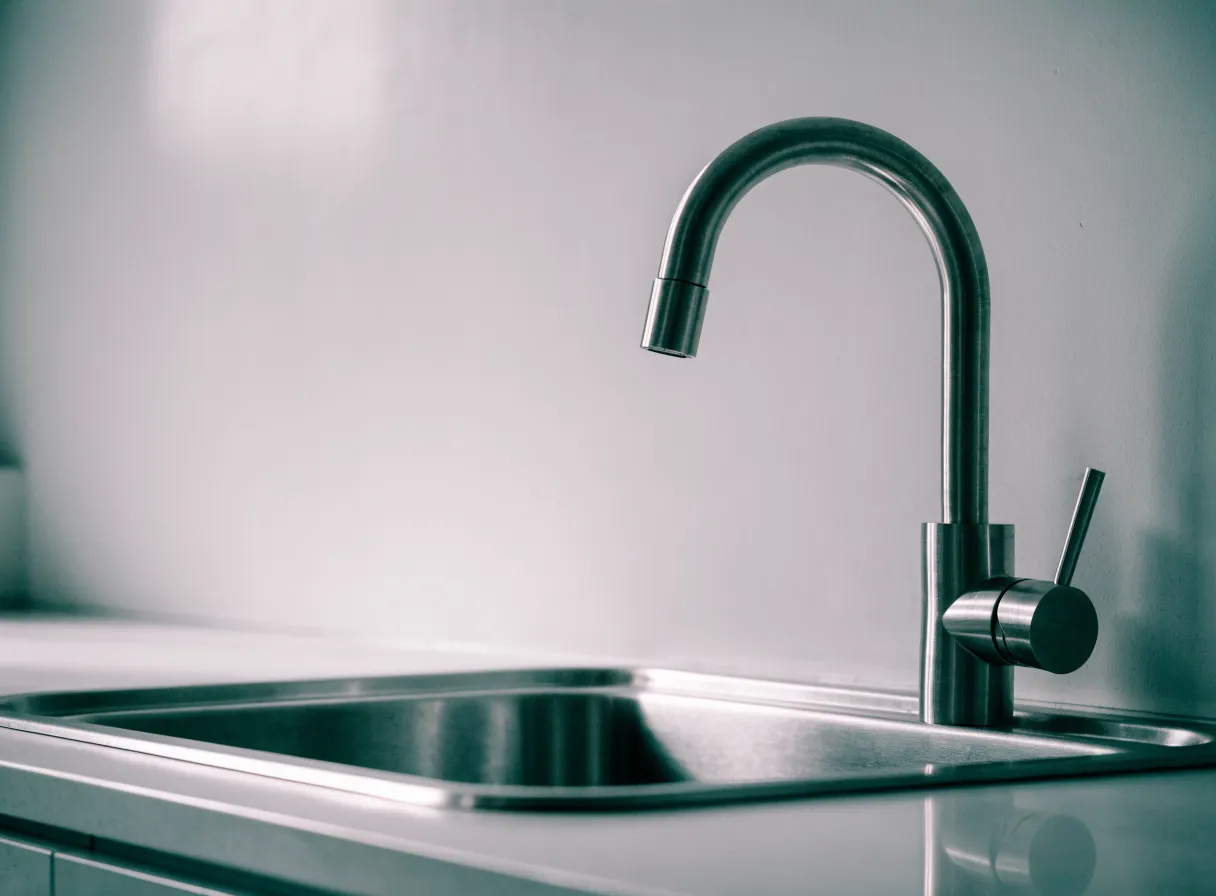 Tall arched brushed steel kitchen faucet over a stainless sink against a plain light wall